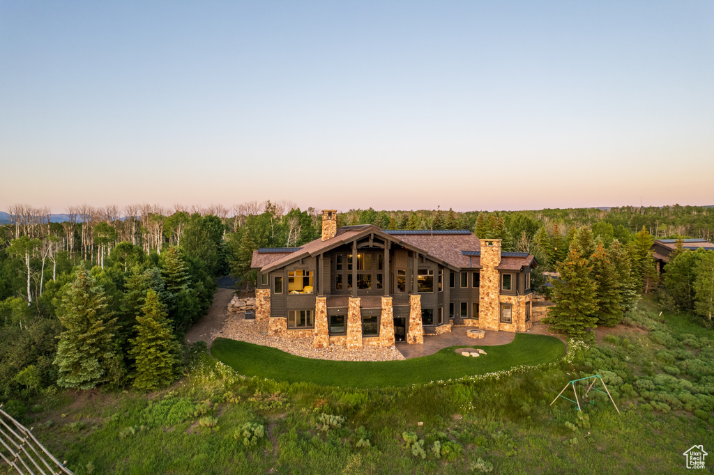 Back of house at dusk featuring a chimney, stone siding, a patio area, and a lawn