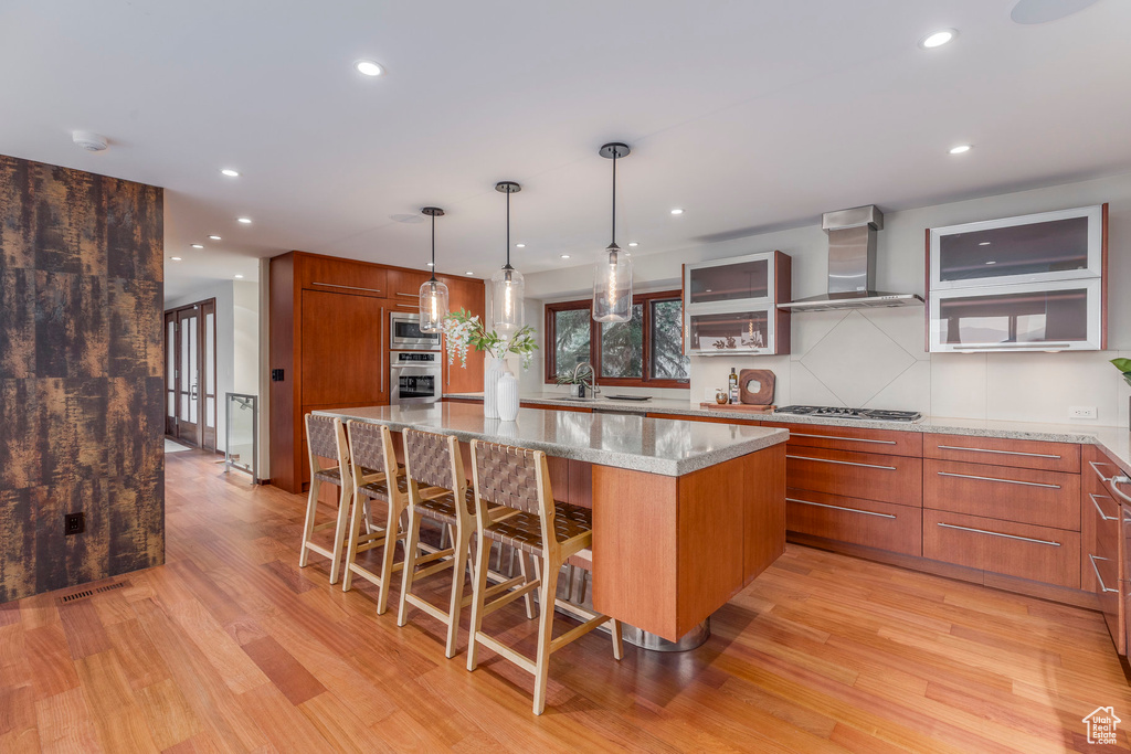 Kitchen featuring wall chimney range hood, appliances with stainless steel finishes, light wood-style flooring, a spacious island, and brown cabinets