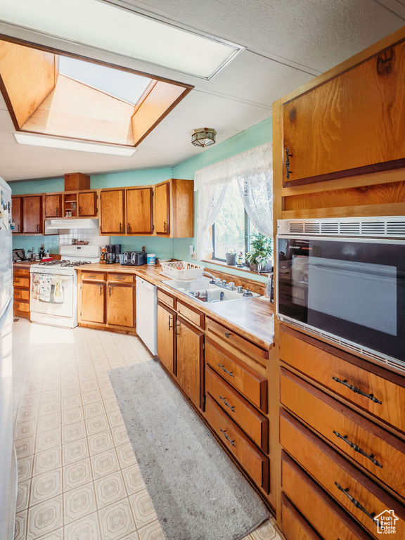 Kitchen with white appliances, a skylight, brown cabinetry, under cabinet range hood, and light countertops