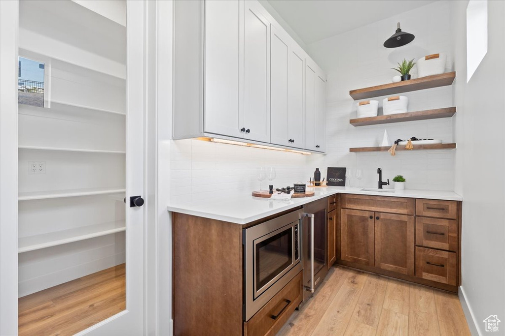 Indoor wet bar featuring stainless steel microwave, light wood-type flooring, decorative backsplash, and beverage cooler