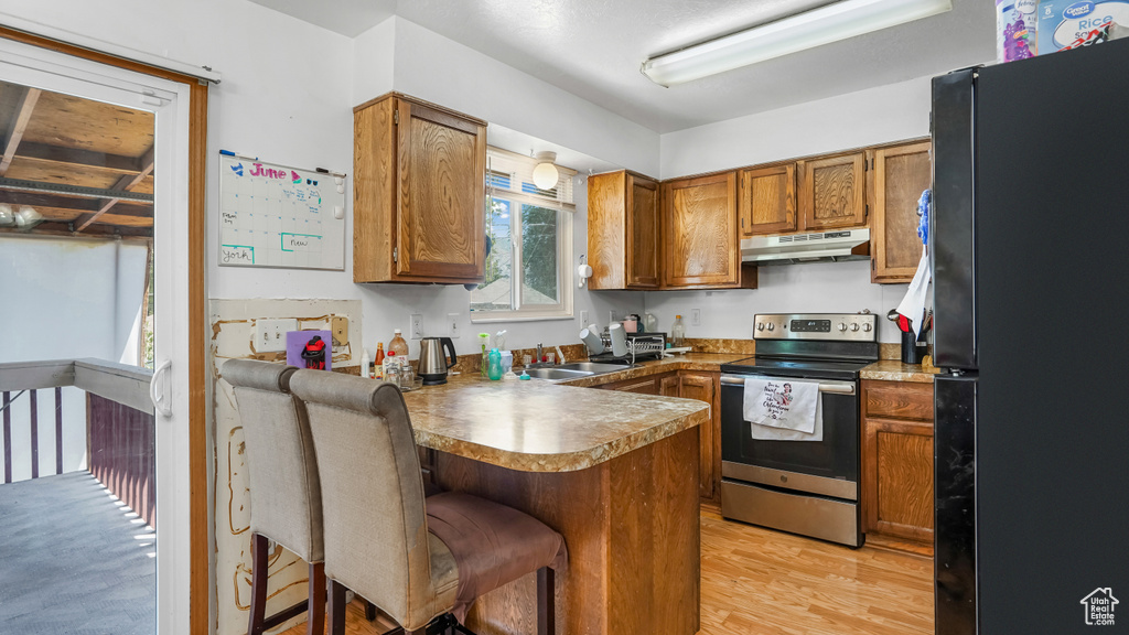 Kitchen with freestanding refrigerator, stainless steel electric range oven, under cabinet range hood, a peninsula, and brown cabinets