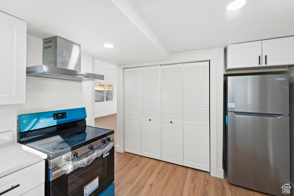 Kitchen with appliances with stainless steel finishes, wall chimney range hood, light wood-type flooring, light countertops, and white cabinets
