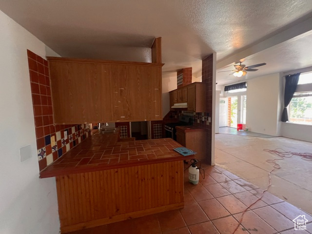Kitchen featuring tile countertops, electric stove, a ceiling fan, under cabinet range hood, and a peninsula