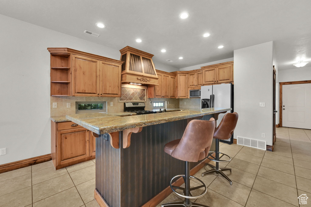 Kitchen with appliances with stainless steel finishes, premium range hood, light tile patterned floors, tasteful backsplash, and a breakfast bar