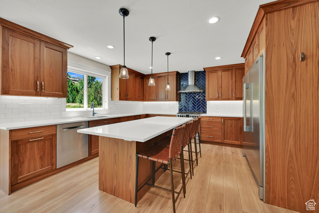 Kitchen with stainless steel appliances, wall chimney exhaust hood, light wood-style flooring, brown cabinets, and a breakfast bar