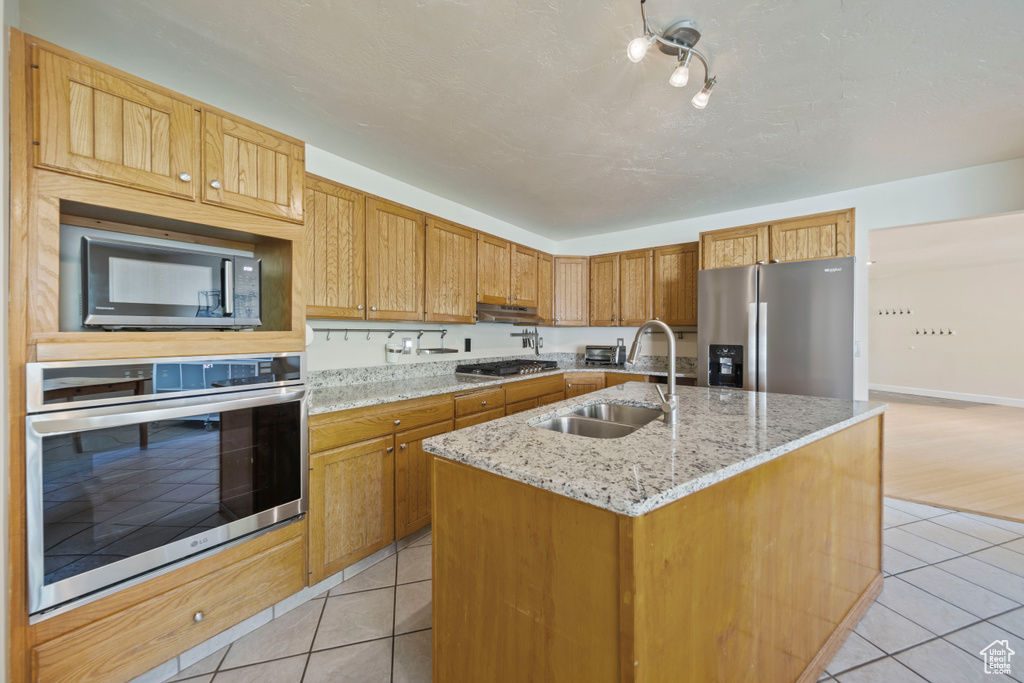 Kitchen featuring appliances with stainless steel finishes, light tile patterned floors, a center island with sink, rail lighting, and light stone counters