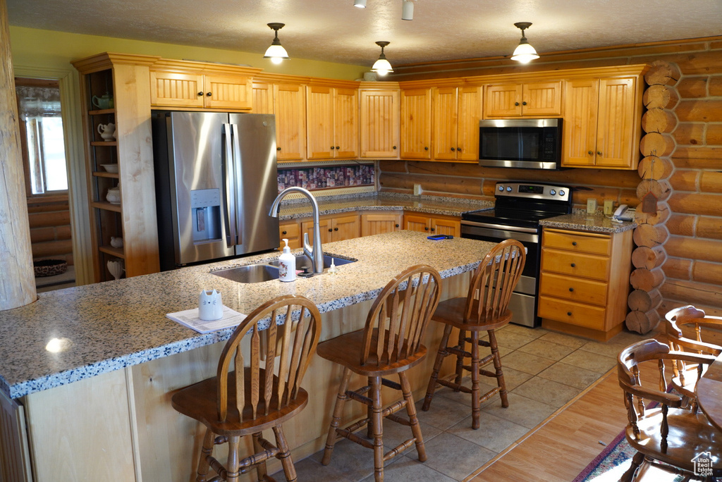 Kitchen with appliances with stainless steel finishes, log walls, a breakfast bar, decorative light fixtures, and light stone counters
