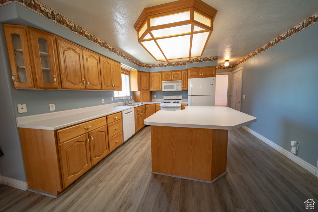 Kitchen with white appliances, wood finished floors, a kitchen island, light countertops, and glass insert cabinets