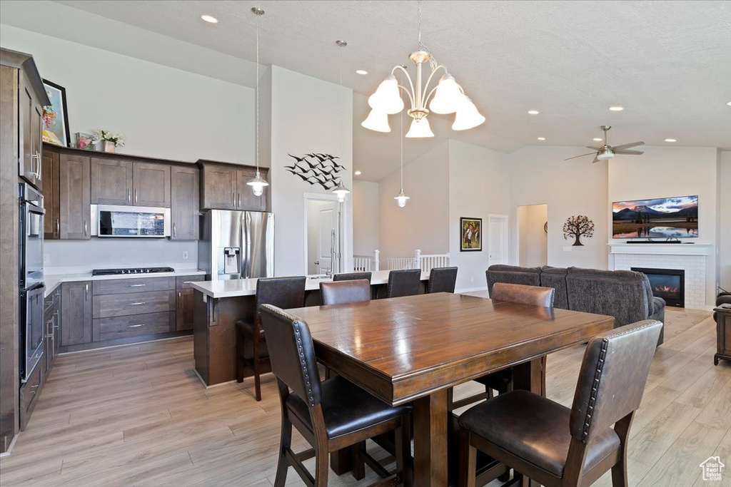 Dining room featuring high vaulted ceiling, a chandelier, light wood-style flooring, a glass covered fireplace, and a ceiling fan