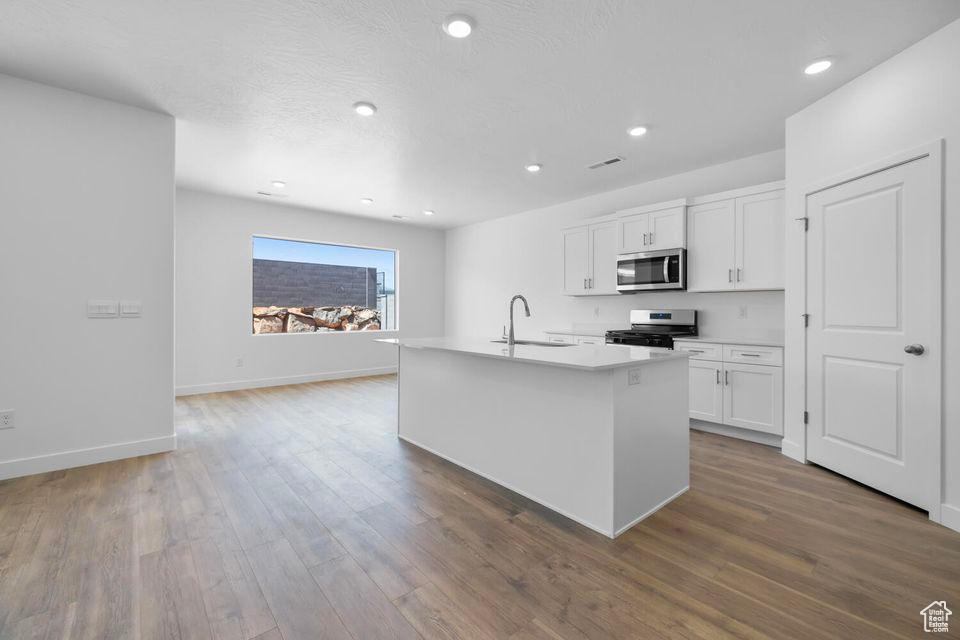 Kitchen with appliances with stainless steel finishes, a kitchen island with sink, white cabinetry, wood finished floors, and light countertops