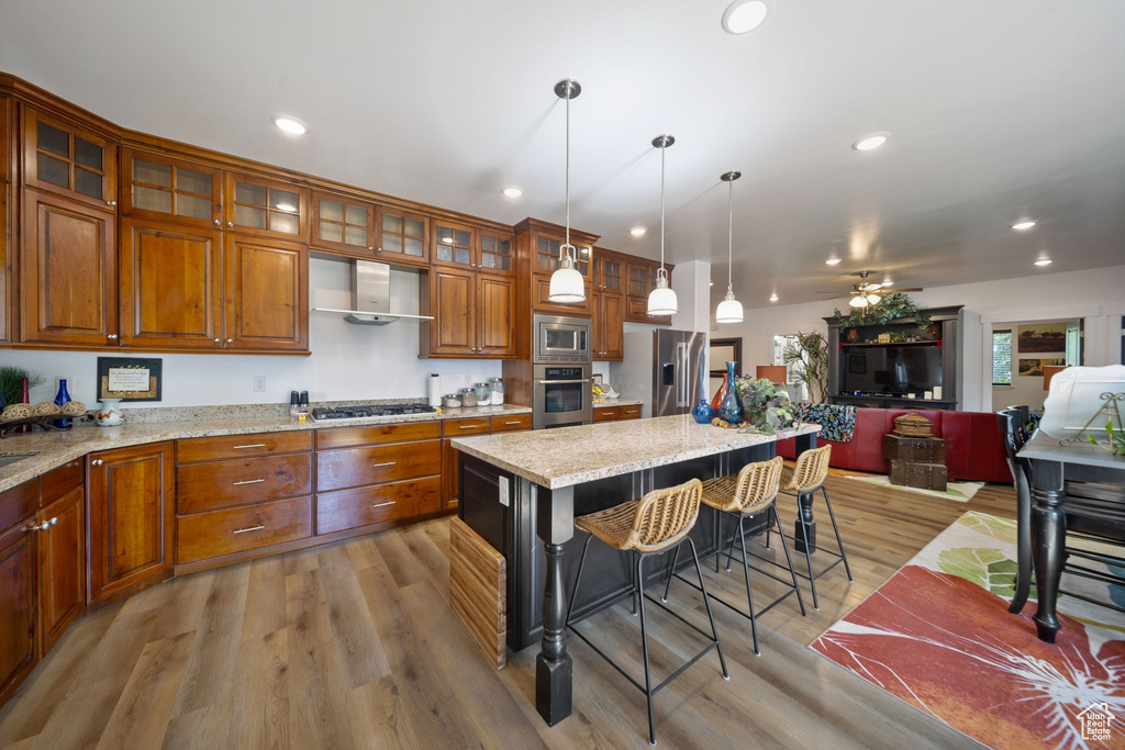 Kitchen featuring appliances with stainless steel finishes, open floor plan, wall chimney range hood, brown cabinets, and a kitchen bar