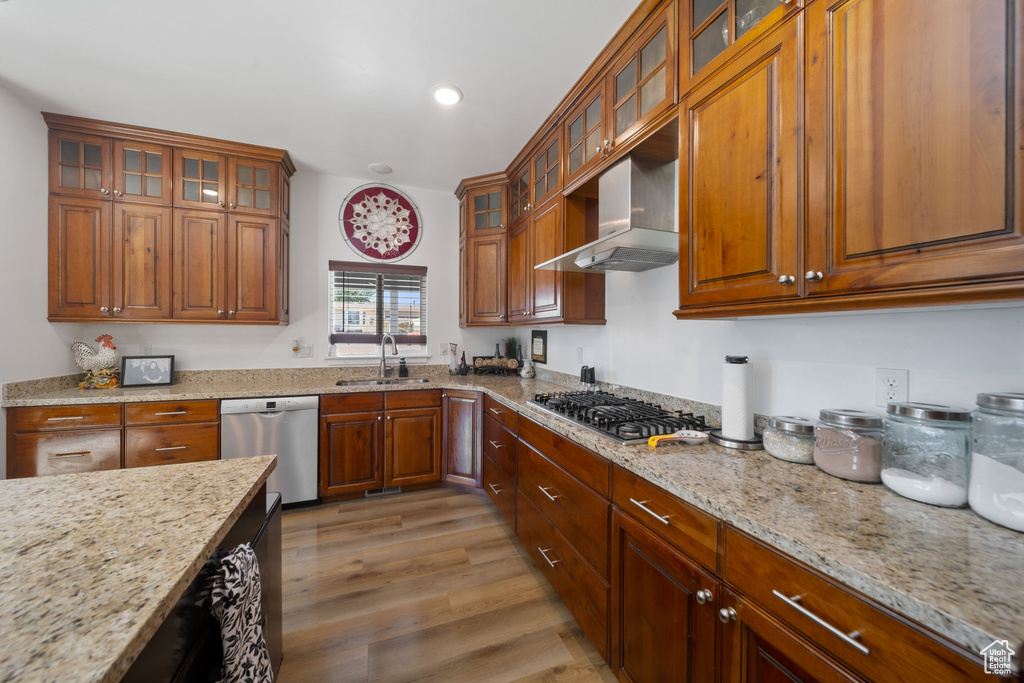 Kitchen with stainless steel appliances, wall chimney range hood, brown cabinetry, light stone counters, and light wood finished floors