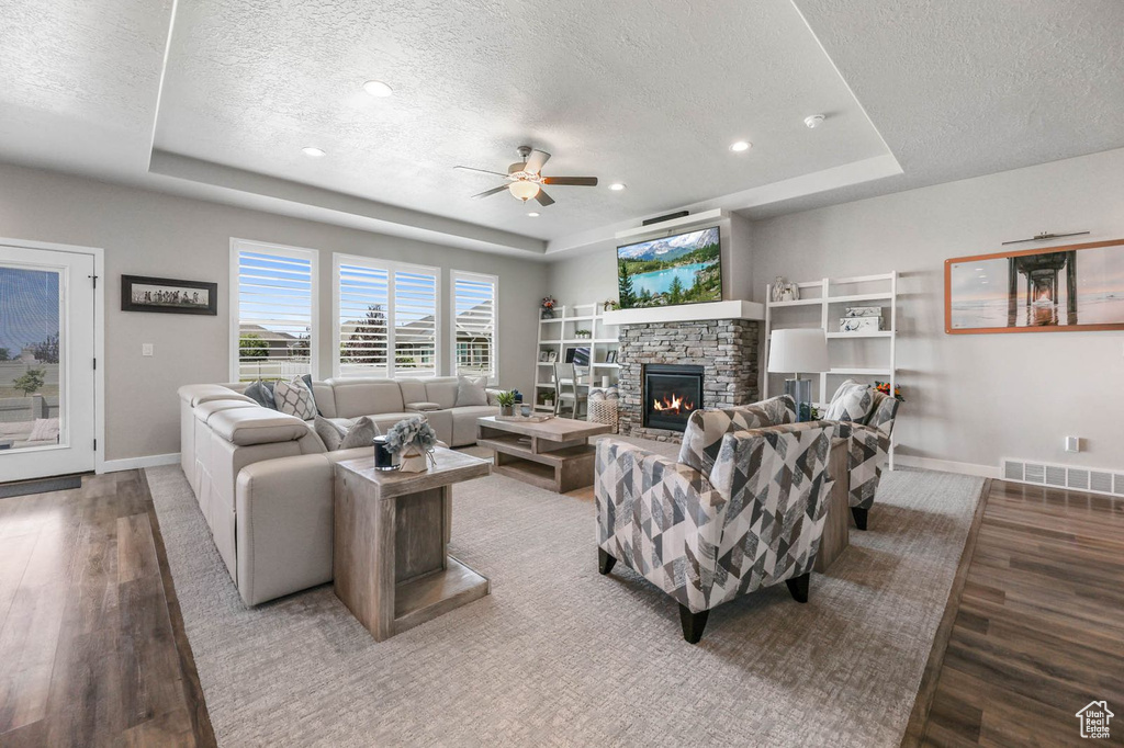 Living area with a raised ceiling, a textured ceiling, wood finished floors, ceiling fan, and a stone fireplace