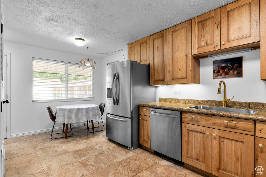 Kitchen with stainless steel appliances, brown cabinetry, dark countertops, a textured ceiling, and decorative light fixtures