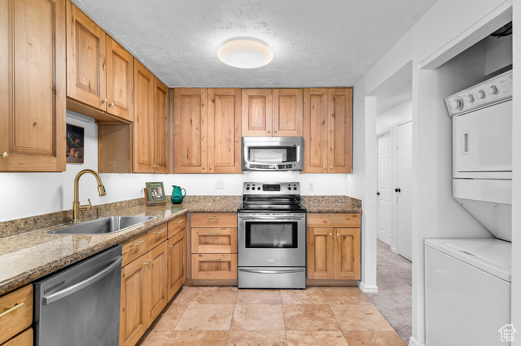 Kitchen featuring stainless steel appliances, stacked washer and clothes dryer, stone countertops, a textured ceiling, and brown cabinetry