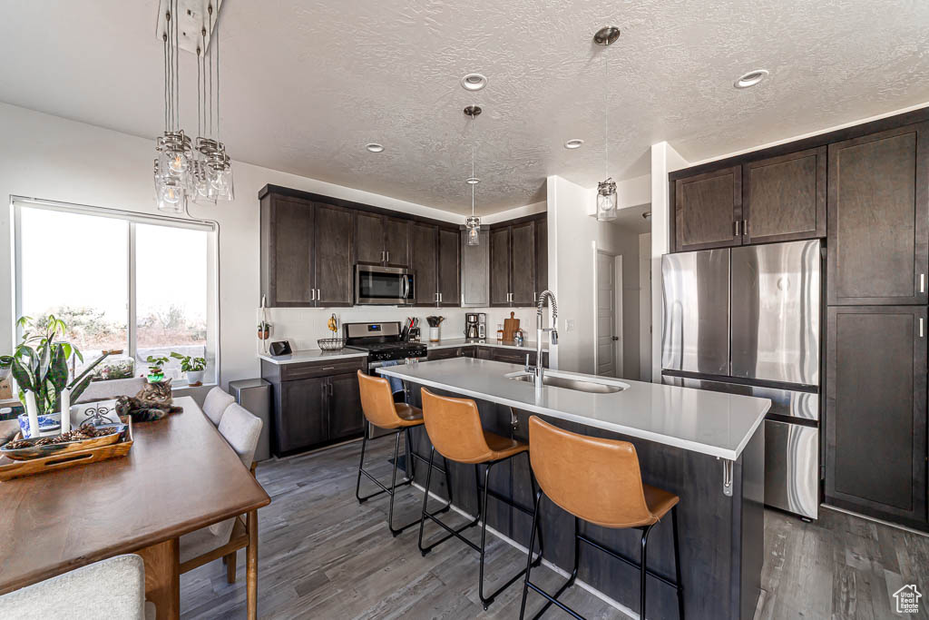 Kitchen with stainless steel appliances, wood finished floors, dark brown cabinetry, a kitchen bar, and a center island with sink