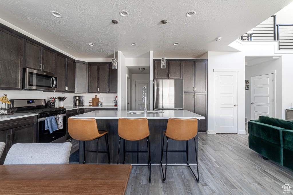 Kitchen with stainless steel appliances, a textured ceiling, dark brown cabinetry, light wood-type flooring, and an island with sink