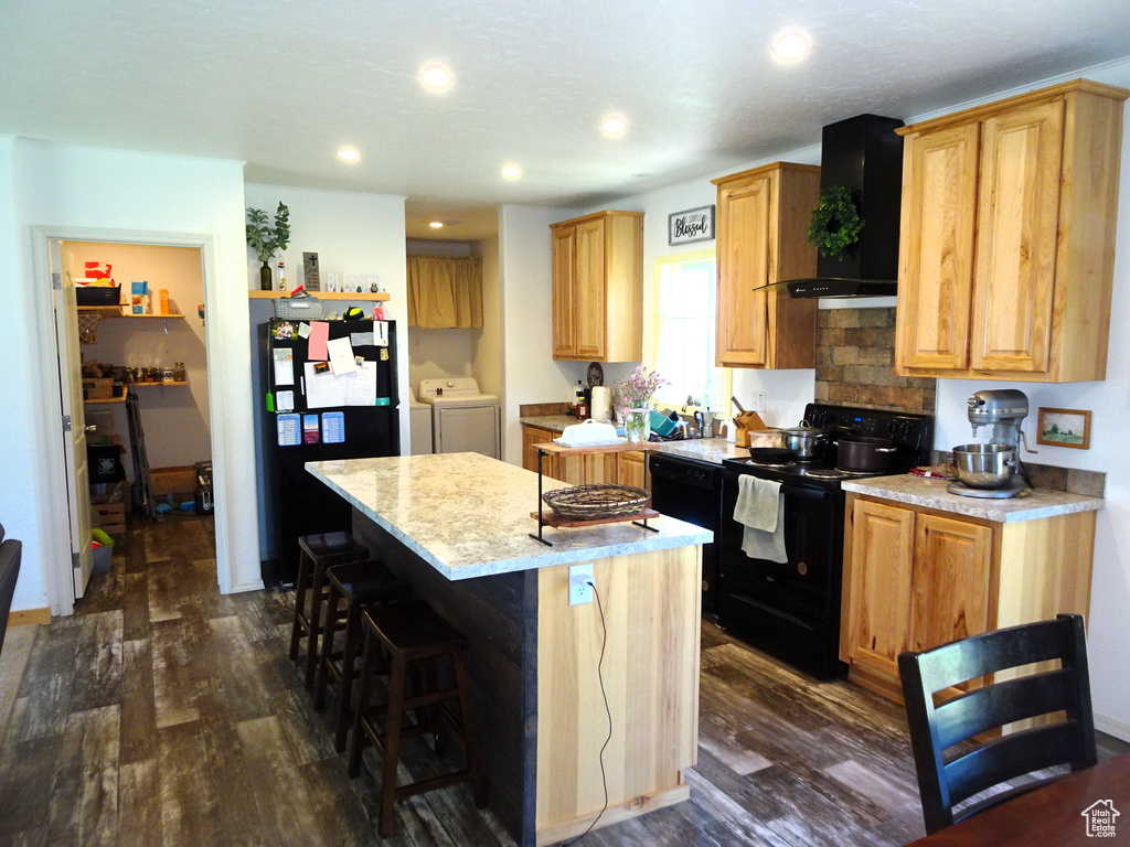 Kitchen with black appliances, wall chimney exhaust hood, independent washer and dryer, a center island, and dark wood-type flooring
