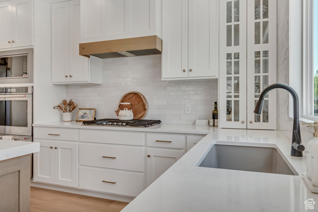 Kitchen with appliances with stainless steel finishes, wall chimney exhaust hood, decorative backsplash, white cabinets, and glass insert cabinets
