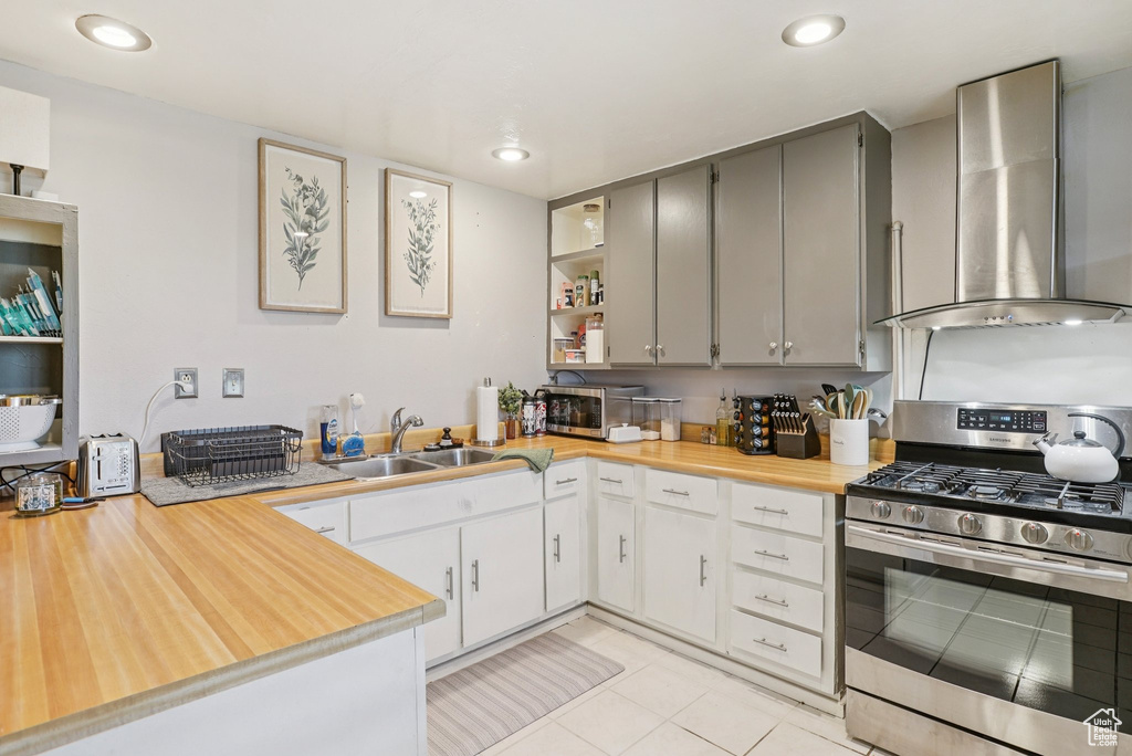 Kitchen with stainless steel gas stove, wall chimney range hood, open shelves, gray cabinets, and light tile patterned floors