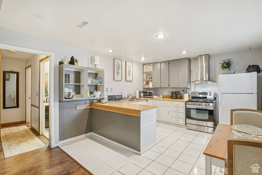 Kitchen featuring wall chimney range hood, stainless steel appliances, a peninsula, wooden counters, and recessed lighting