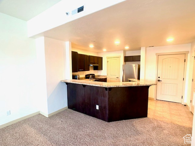 Kitchen with freestanding refrigerator, dark brown cabinets, a peninsula, recessed lighting, and light stone countertops