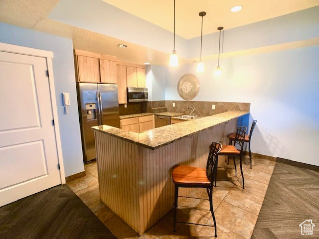 Kitchen with appliances with stainless steel finishes, a peninsula, a kitchen bar, light brown cabinetry, and stone countertops