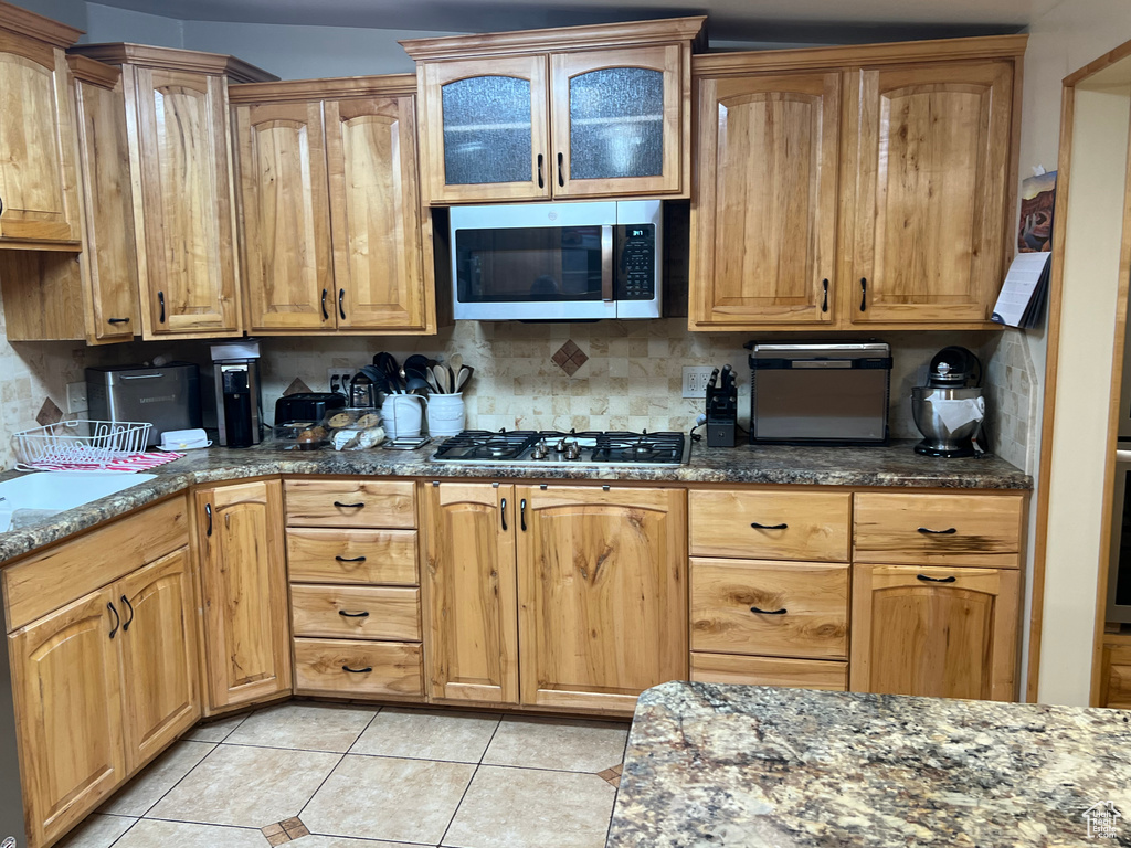 Kitchen with appliances with stainless steel finishes, dark stone countertops, light tile patterned flooring, and glass insert cabinets