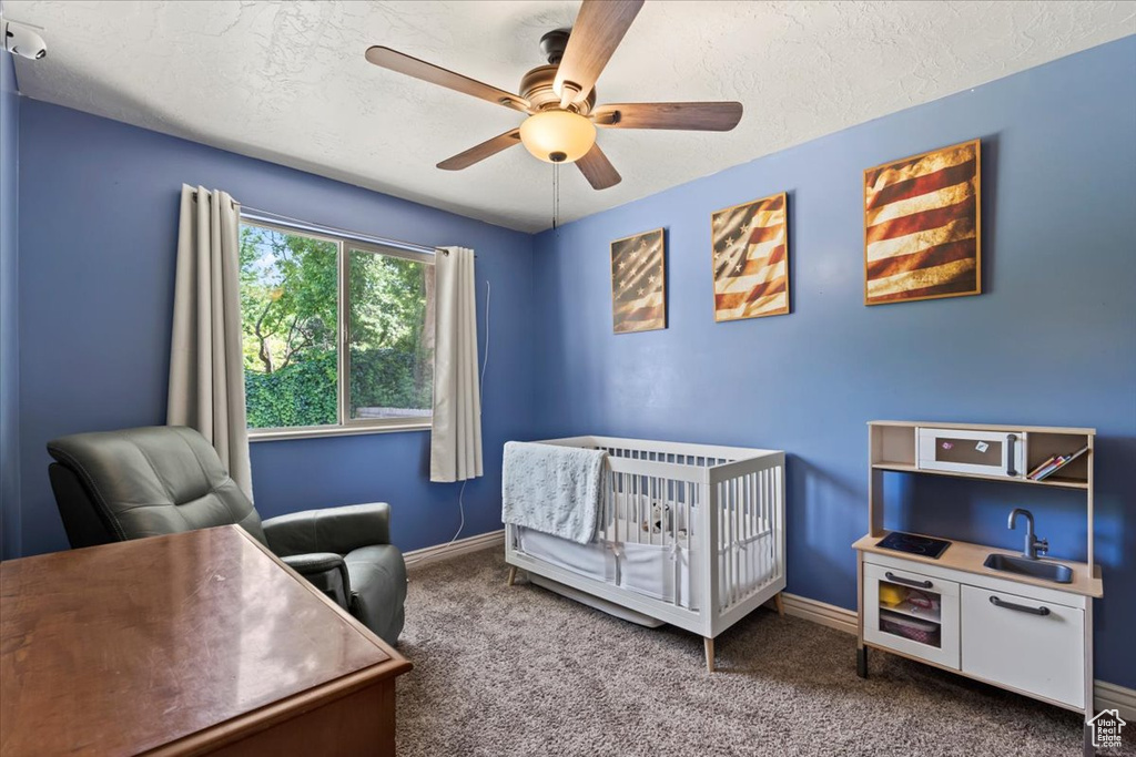 Carpeted bedroom featuring a textured ceiling, ceiling fan, a desk, and a crib