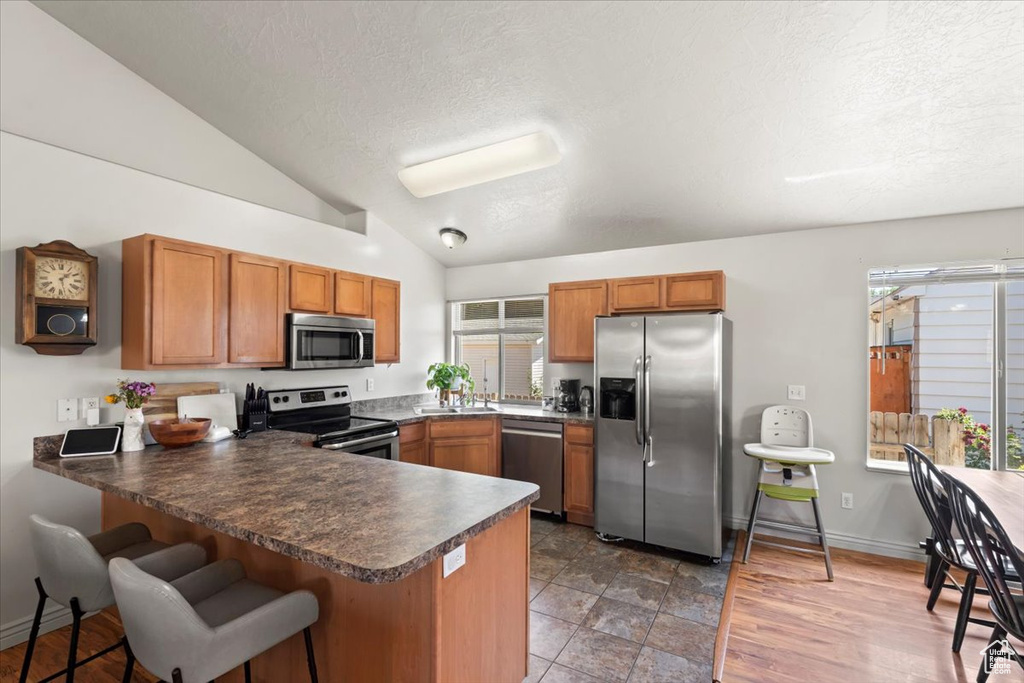 Kitchen with stainless steel appliances, a peninsula, lofted ceiling, brown cabinets, and dark countertops