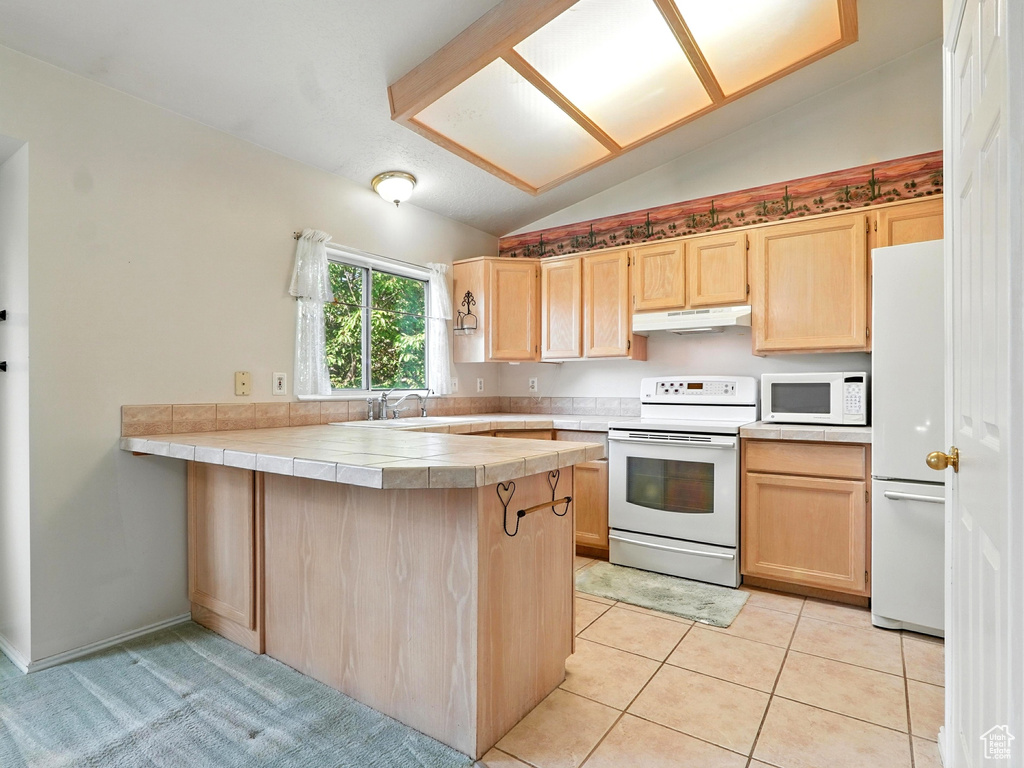 Kitchen with white appliances, under cabinet range hood, light brown cabinetry, lofted ceiling, and a peninsula