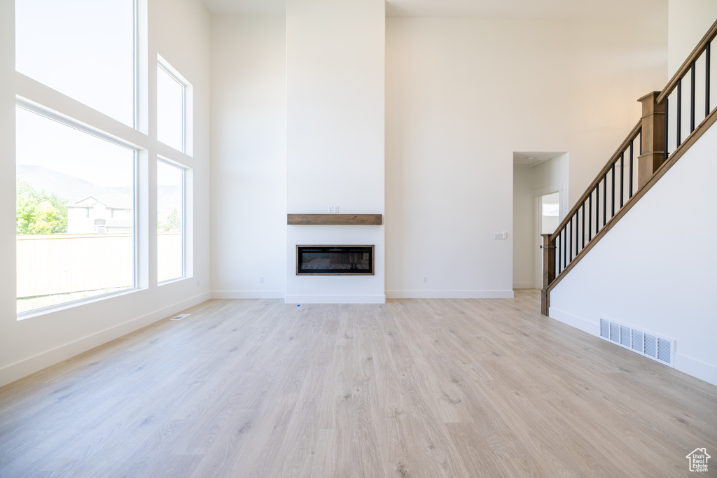 Unfurnished living room featuring a high ceiling, stairs, light wood-style flooring, and a glass covered fireplace