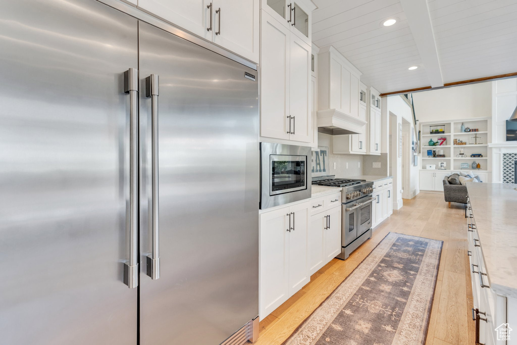 Kitchen with built in appliances, beamed ceiling, white cabinets, glass insert cabinets, and light wood-style flooring