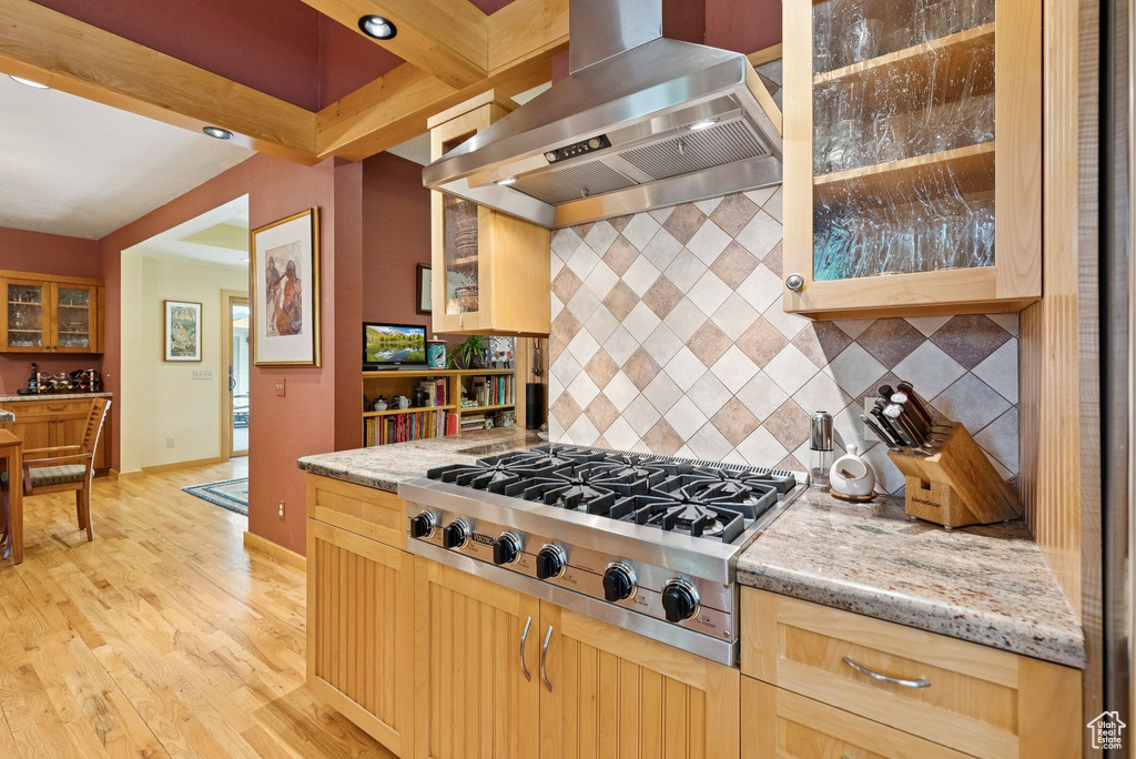 Kitchen featuring wall chimney range hood, stainless steel gas stovetop, decorative backsplash, light wood finished floors, and light stone countertops