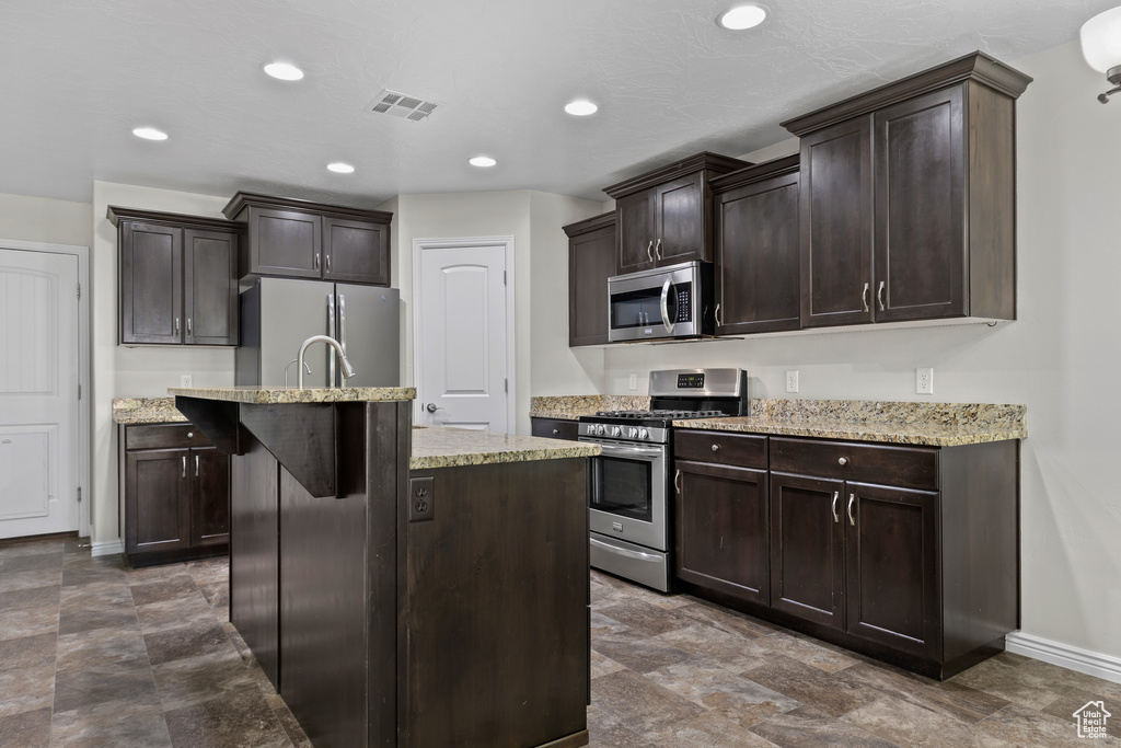 Kitchen featuring appliances with stainless steel finishes, dark brown cabinetry, a center island with sink, light stone counters, and recessed lighting