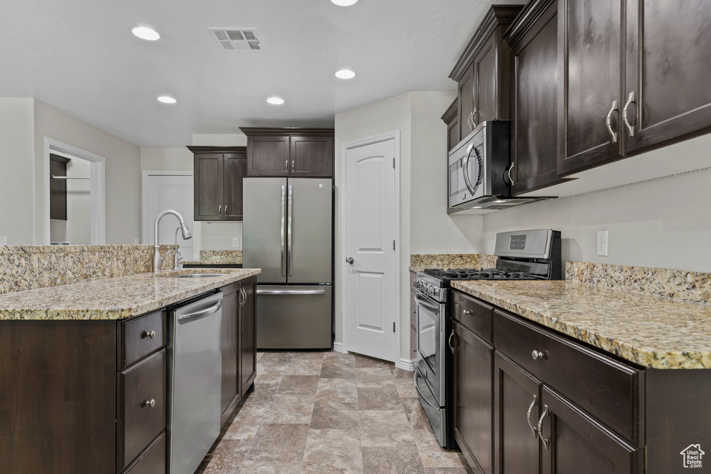 Kitchen featuring appliances with stainless steel finishes, dark brown cabinetry, light stone counters, recessed lighting, and an island with sink