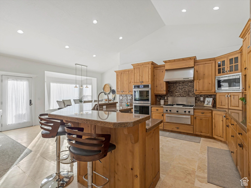 Kitchen featuring appliances with stainless steel finishes, backsplash, a breakfast bar, a center island with sink, and custom exhaust hood