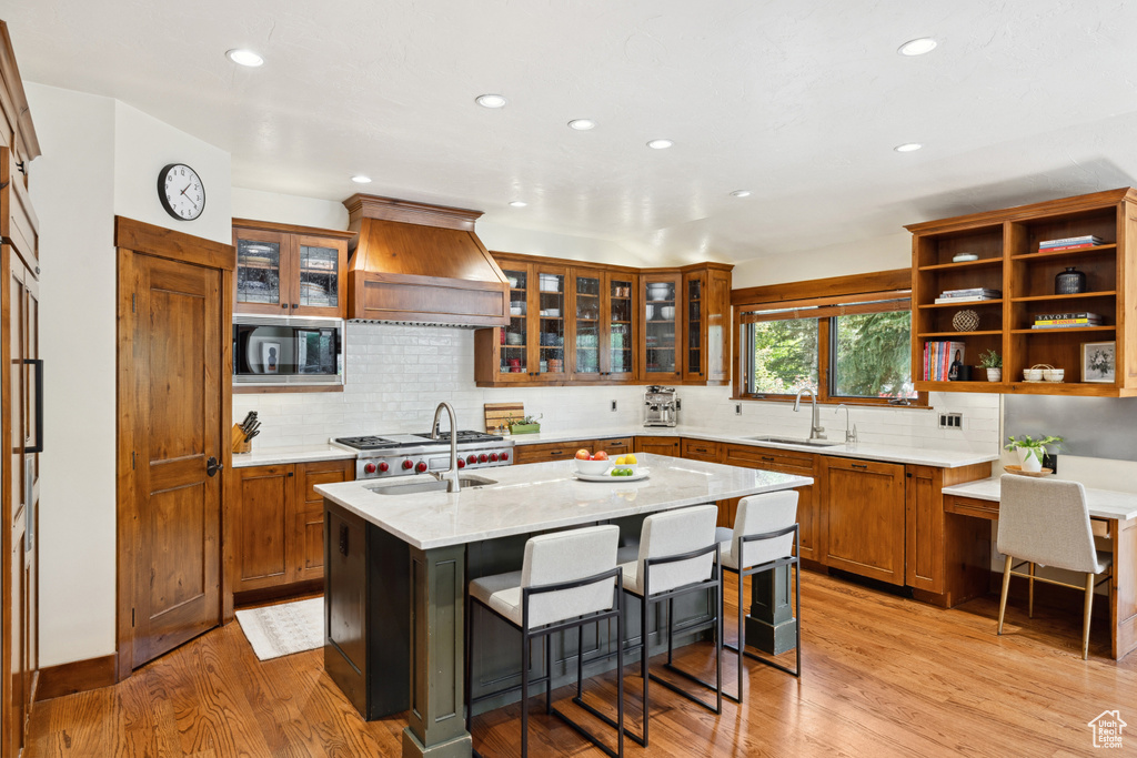 Kitchen featuring custom range hood, stainless steel microwave, recessed lighting, a kitchen island with sink, and light wood finished floors