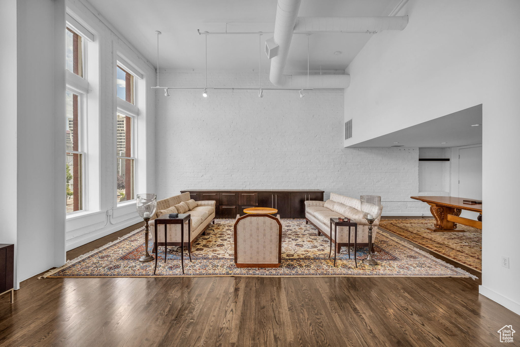 Living room with rail lighting, wood finished floors, plenty of natural light, brick wall, and a towering ceiling