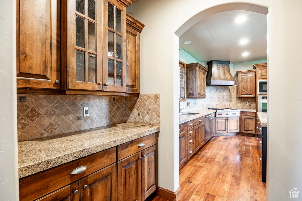 Kitchen with custom exhaust hood, light wood-style flooring, arched walkways, tasteful backsplash, and stainless steel appliances
