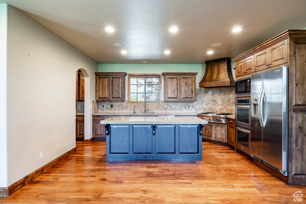 Kitchen featuring stainless steel appliances, custom range hood, arched walkways, light wood-style floors, and backsplash