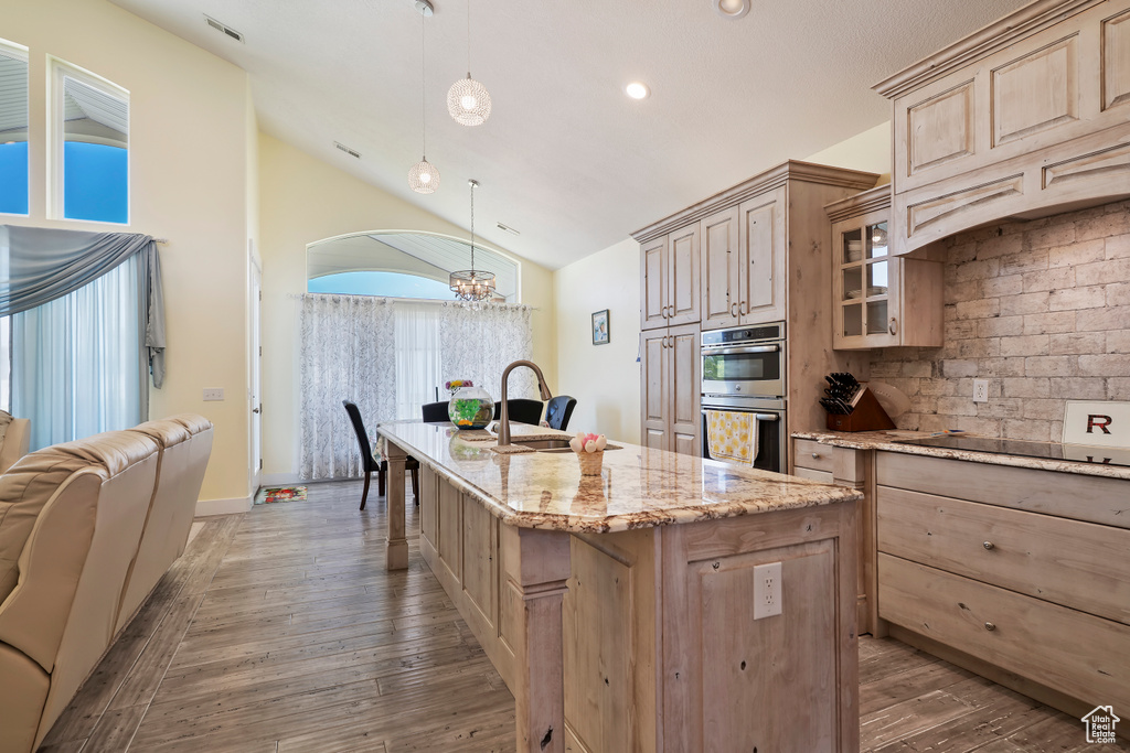 Kitchen with light brown cabinets, double oven, black electric cooktop, a center island with sink, and vaulted ceiling