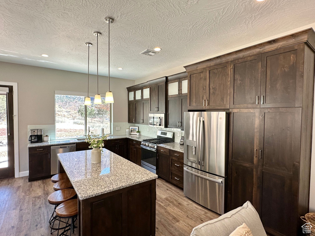 Kitchen with appliances with stainless steel finishes, dark brown cabinets, glass insert cabinets, a breakfast bar area, and light wood-style flooring