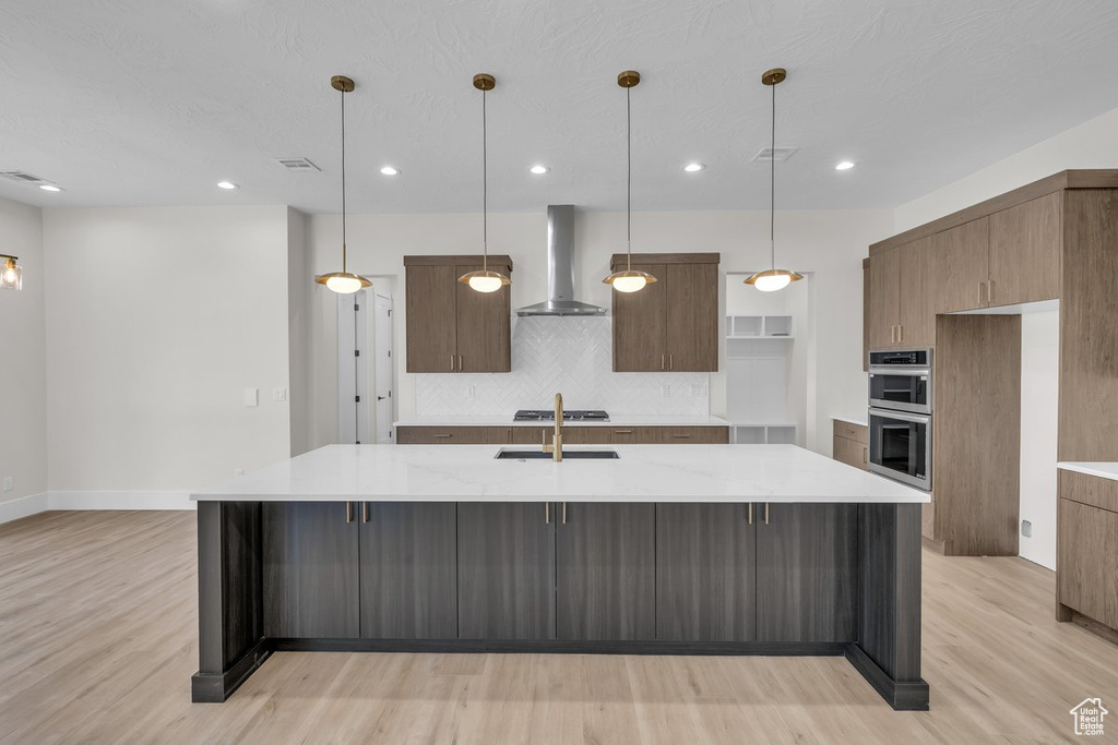 Kitchen with wall chimney range hood, double oven, light wood finished floors, decorative backsplash, and a large island with sink