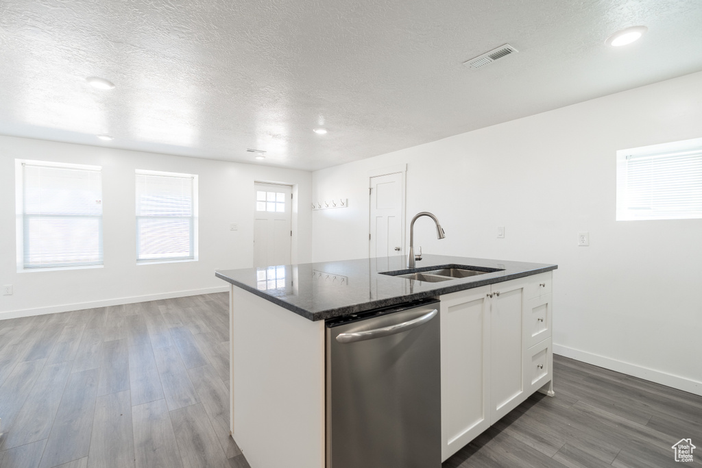 Kitchen featuring stainless steel dishwasher, wood finished floors, a center island with sink, a textured ceiling, and white cabinets
