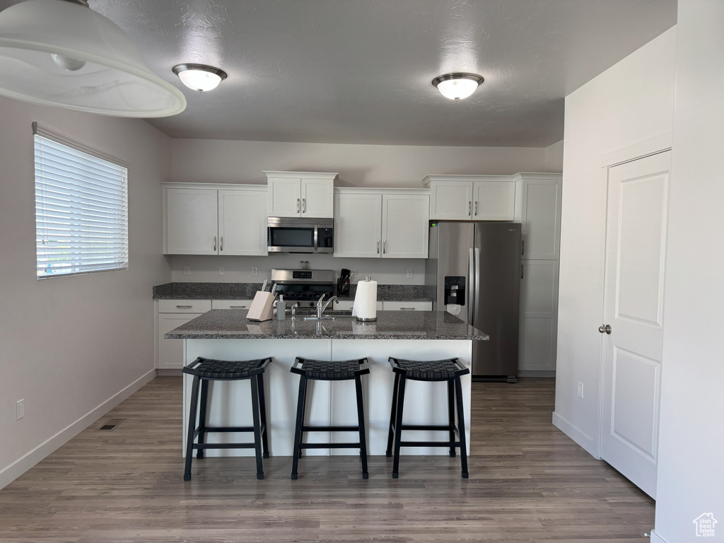 Kitchen featuring stainless steel appliances, a breakfast bar area, white cabinets, an island with sink, and wood finished floors