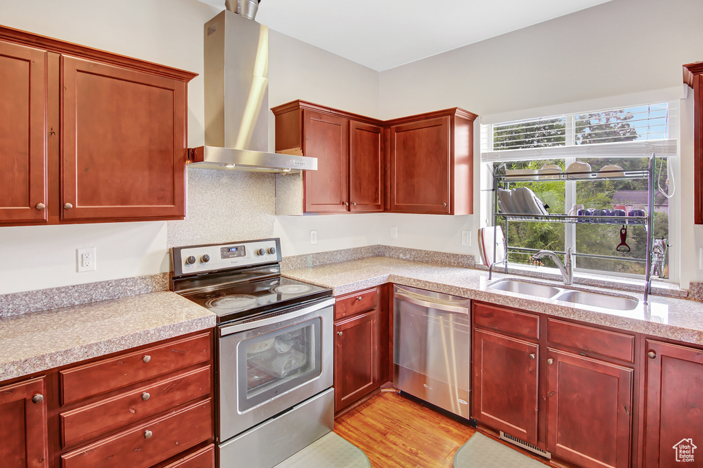 Kitchen with appliances with stainless steel finishes, wall chimney range hood, reddish brown cabinets, light countertops, and light wood-style floors