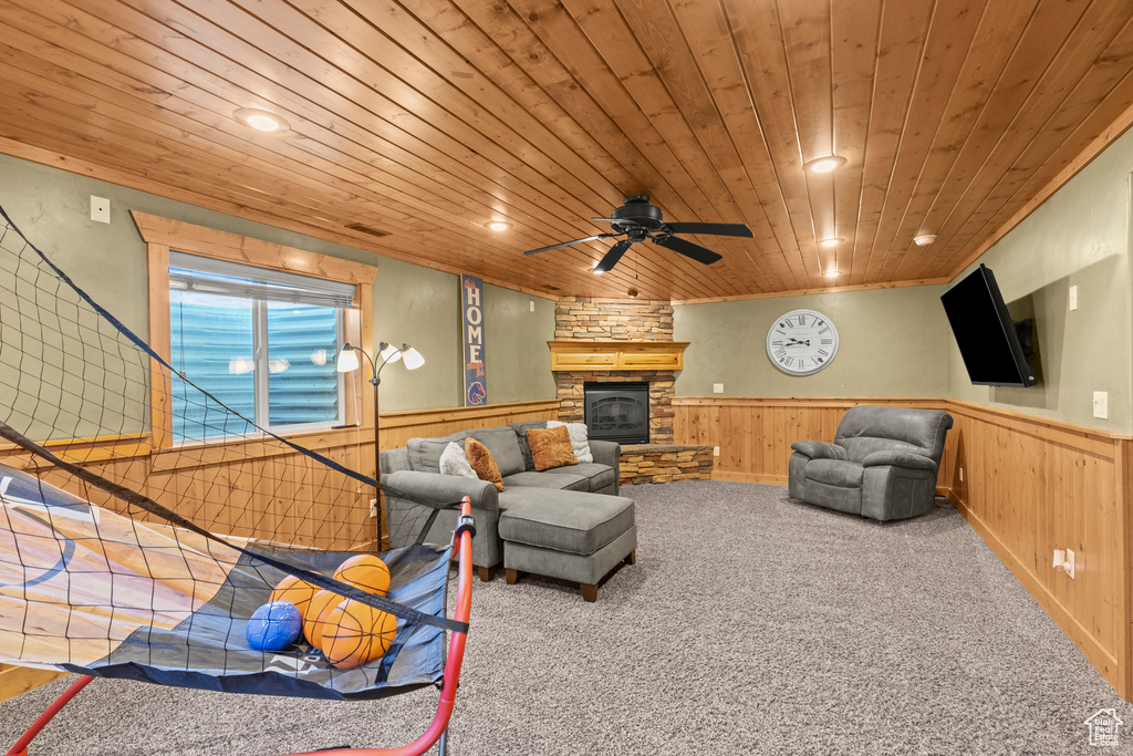 Carpeted living area featuring a wainscoted wall, wooden ceiling, a stone fireplace, and a ceiling fan
