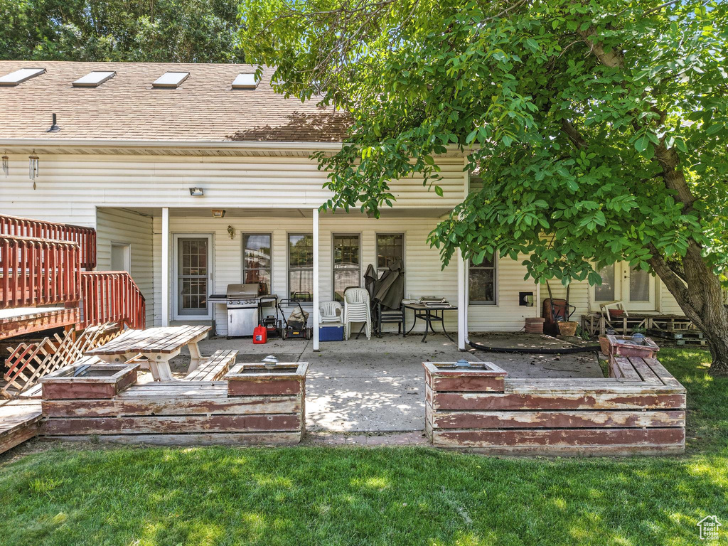 Back of property featuring a wooden deck, roof with shingles, a patio area, and a lawn