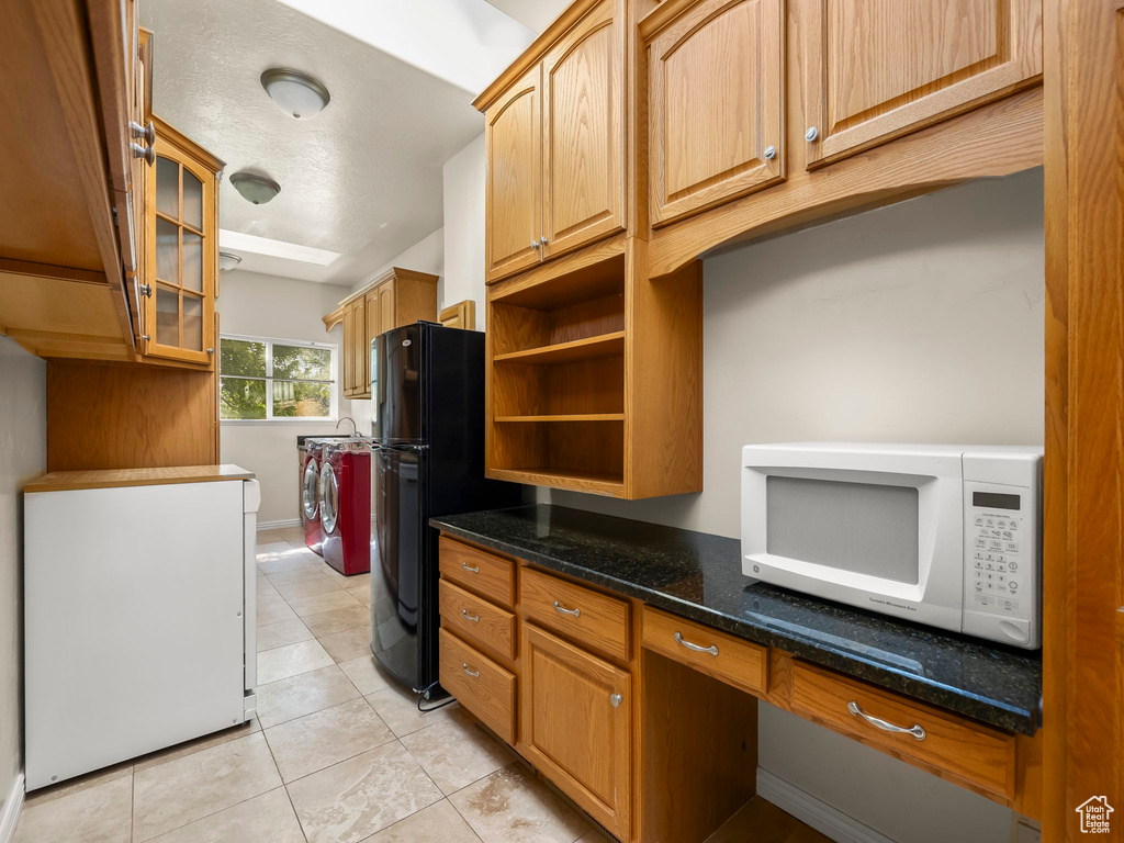 Kitchen with white microwave, washing machine and dryer, freestanding refrigerator, light tile patterned floors, and dark stone counters